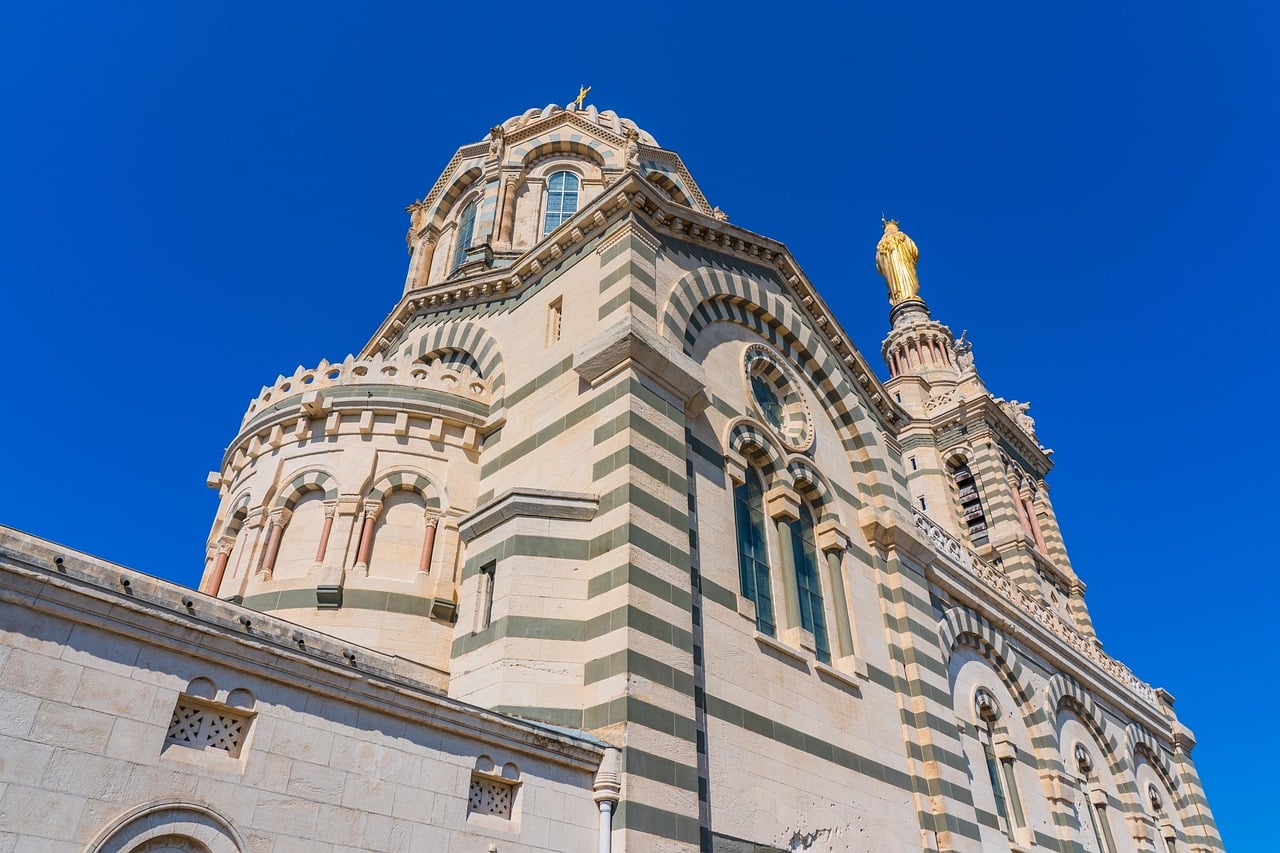 마르세유 노트르담 드 라 가르드 성당, 푸른 하늘 아래 줄무늬 대리석 외벽과 종탑 꼭대기의 황금빛 성모 마리아 상이 돋보이는 웅장한 모습입니다.