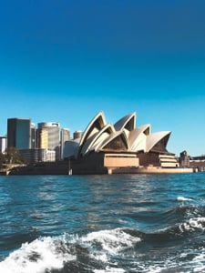 시드니 오페라하우스, 쾌청한 하늘 아래 푸른 바다 물결 너머로 보이는 웅장한 하얀 돛 모양의 건축물과 도심 스카이라인 풍경입니다.