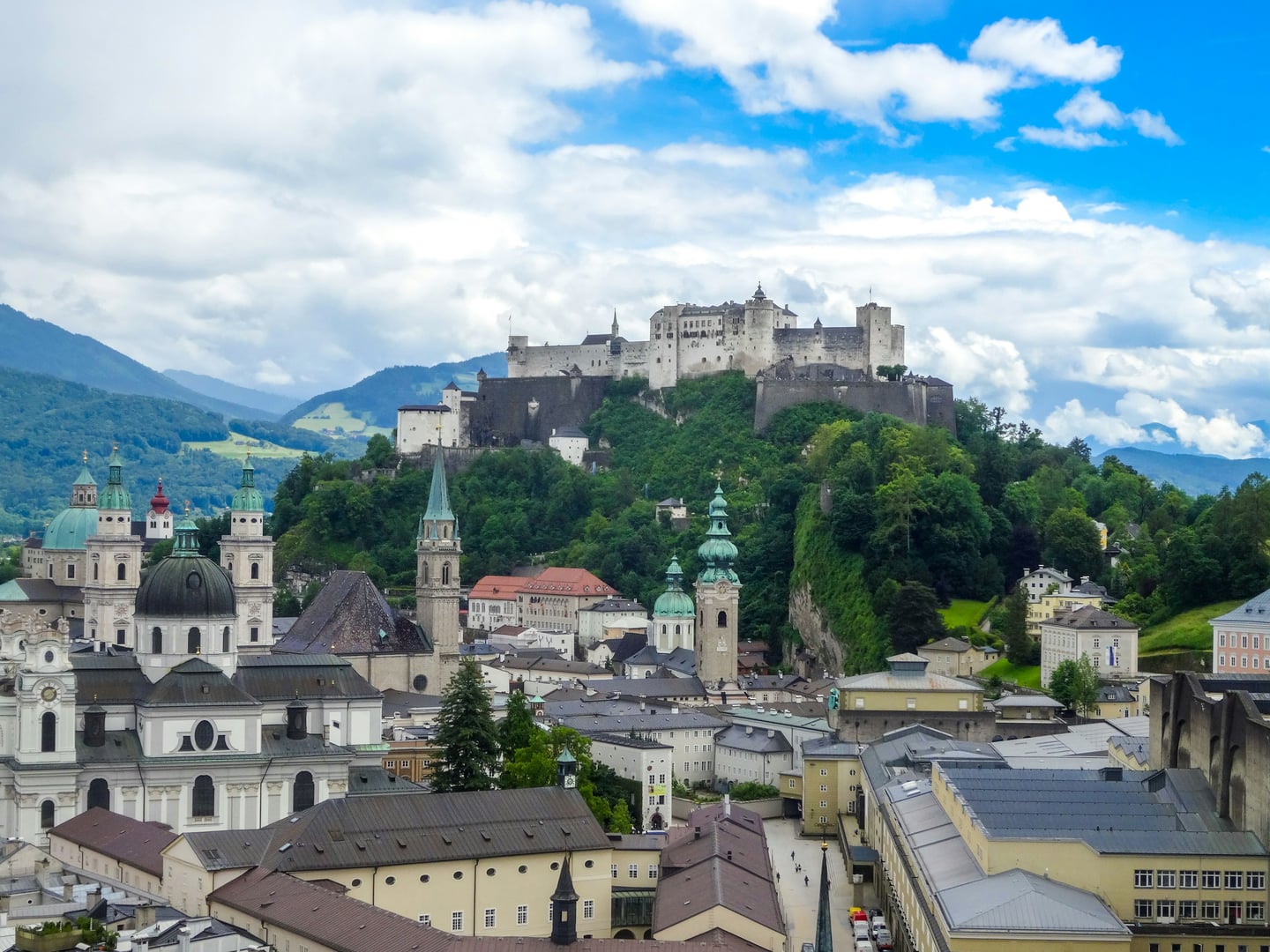 잘츠부르크 호엔잘츠부르크 성 전망, 푸른 하늘 아래 구시가지의 고풍스러운 성당 돔과 언덕 위 웅장한 요새가 어우러진 풍경입니다.