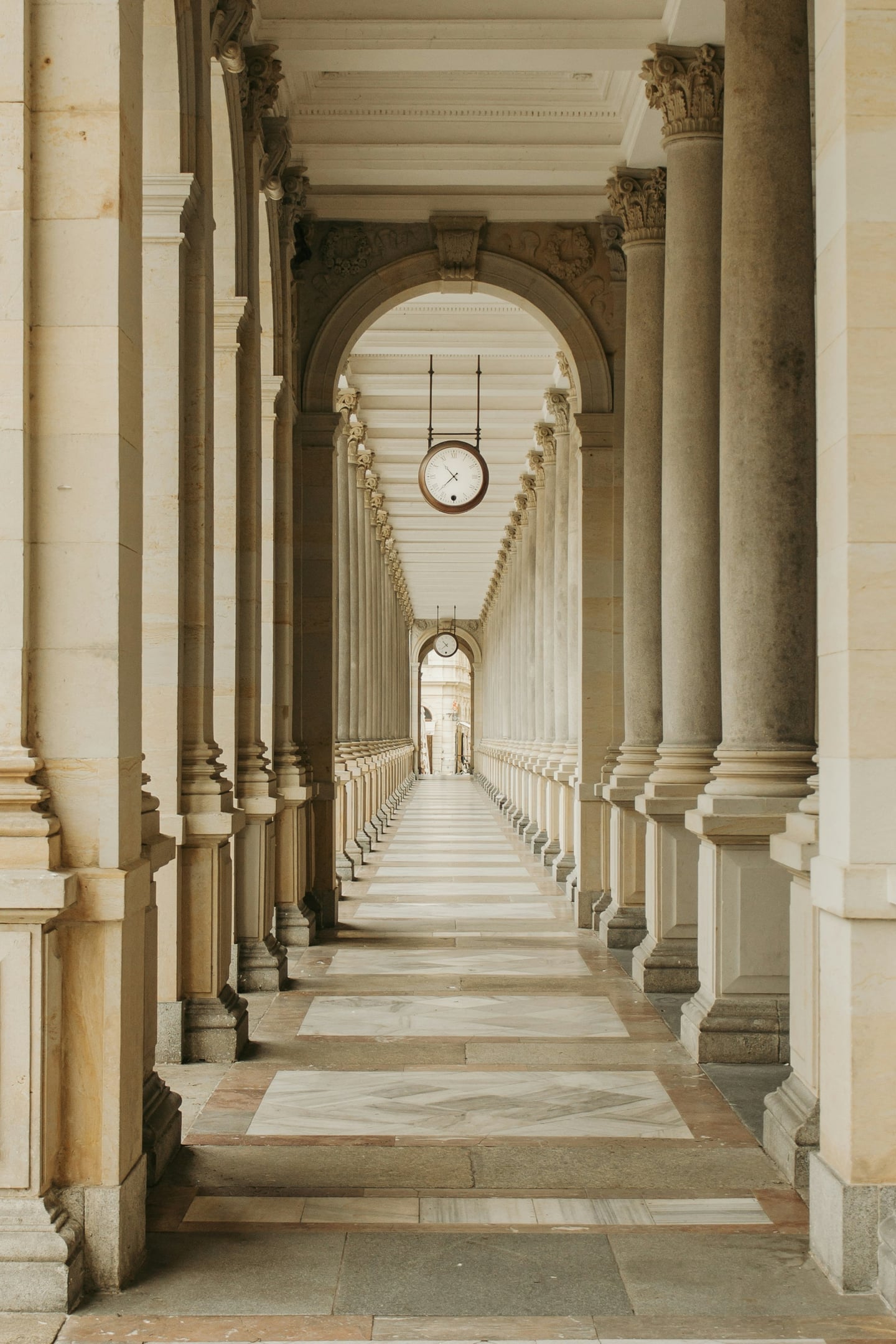 카를로비바리 믈린스카 콜로나다, 웅장한 석조 기둥들이 끝없이 이어지는 회랑 천장에 고풍스러운 시계가 매달려 있는 대칭적인 건축미.