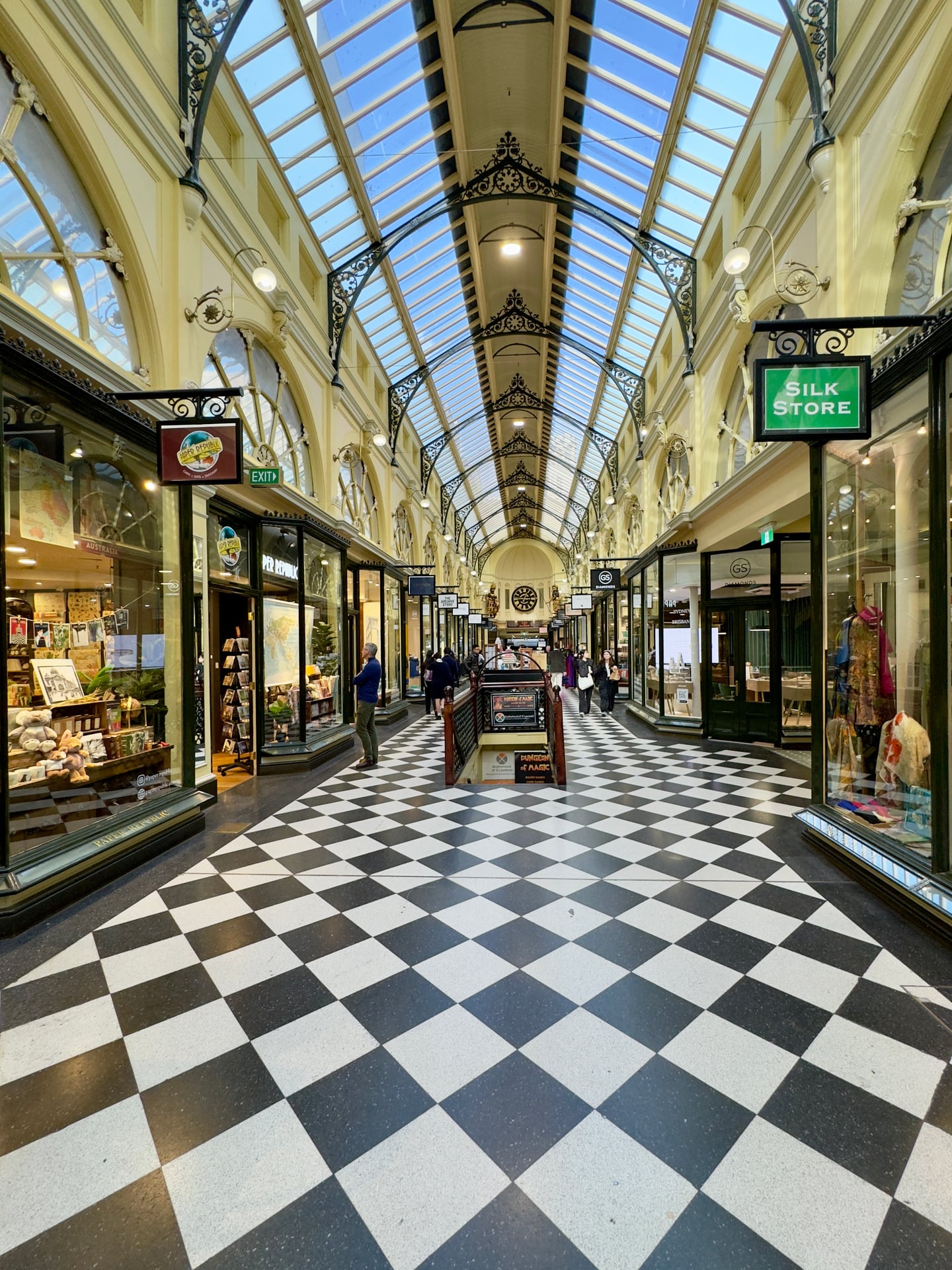 멜버른 로얄 아케이드, 화려한 유리 천장 아래 펼쳐진 흑백 체크무늬 바닥과 고전적인 상점들이 어우러진 고풍스러운 내부 전경입니다.