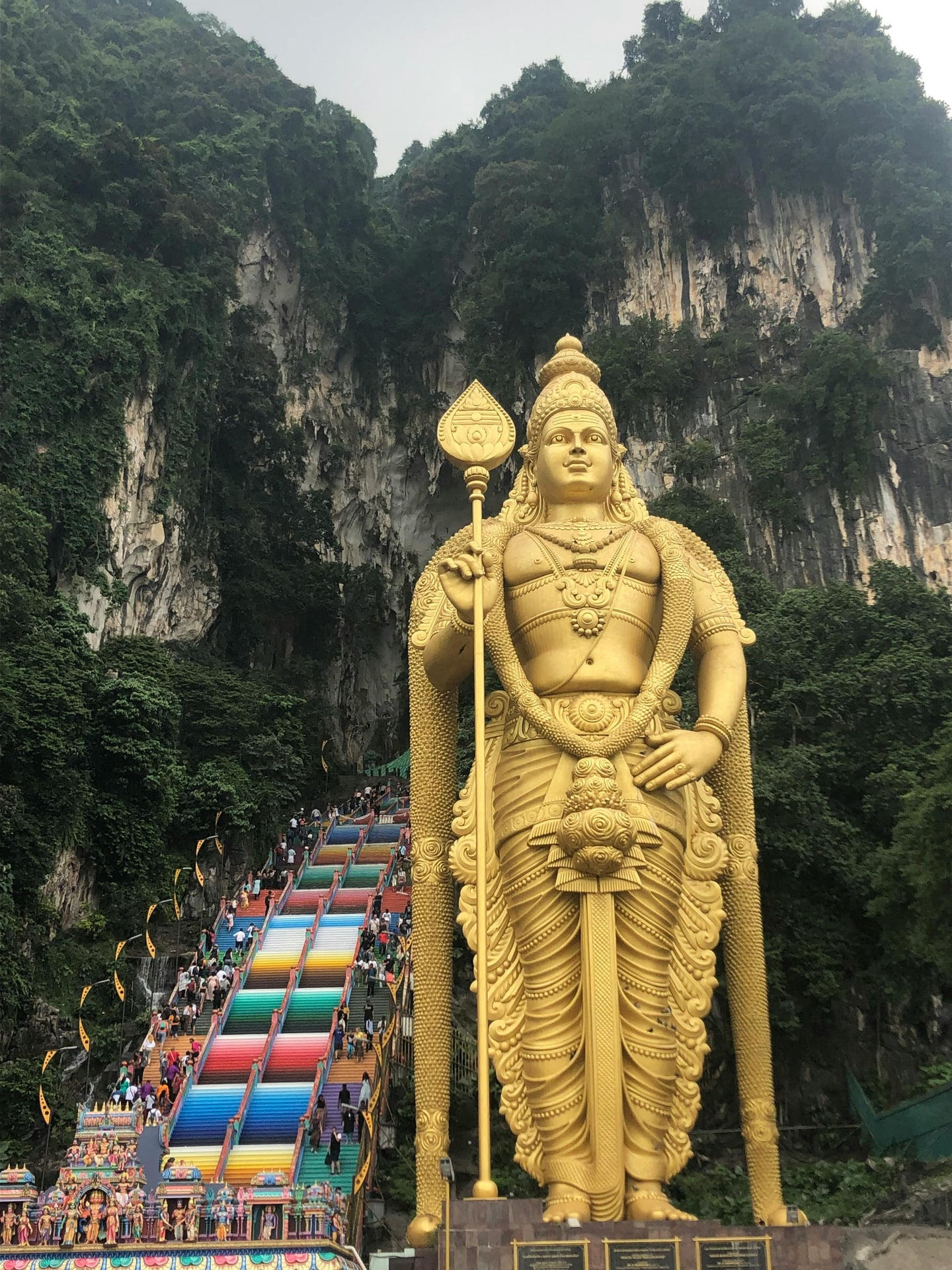 쿠알라룸푸르 바투동굴, 거대한 황금빛 무루간 신상과 가파른 무지개색 계단이 웅장한 석회암 절벽과 어우러진 풍경입니다.