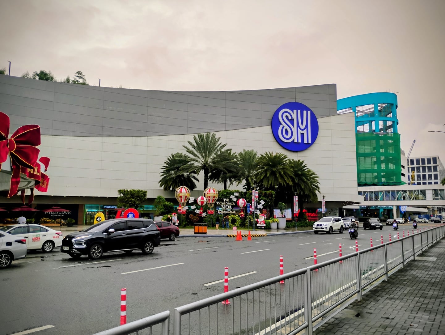 마닐라 SM 몰 오브 아시아, 대형 로고가 선명한 현대적인 쇼핑몰 외관과 야자수가 어우러진 활기찬 도로 풍경입니다.