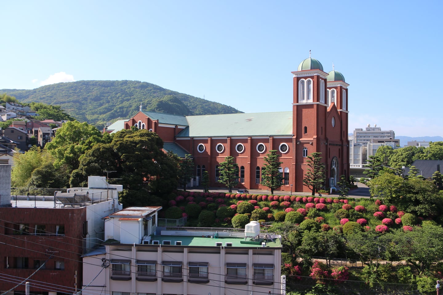 나가사키 우라카미 천주당, 푸른 산을 배경으로 붉은 벽돌 성당과 분홍색 꽃이 만발한 정원이 어우러진 풍경입니다.