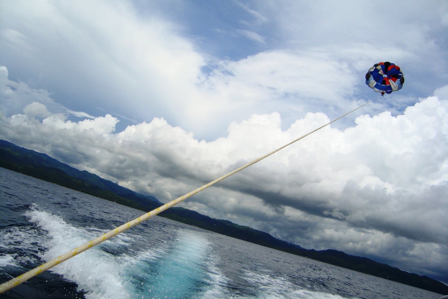 범용 파라세일링, 푸른 바다 위 뭉게구름이 가득한 하늘을 배경으로 화려한 낙하산이 보트에 연결되어 높이 날아오르는 역동적인 모습입니다.
