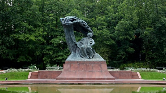 바르샤바 쇼팽 공원, 울창한 숲을 배경으로 연못 앞에 세워진 거대한 프레데리크 쇼팽 기념비와 평화로운 공원 전경입니다.