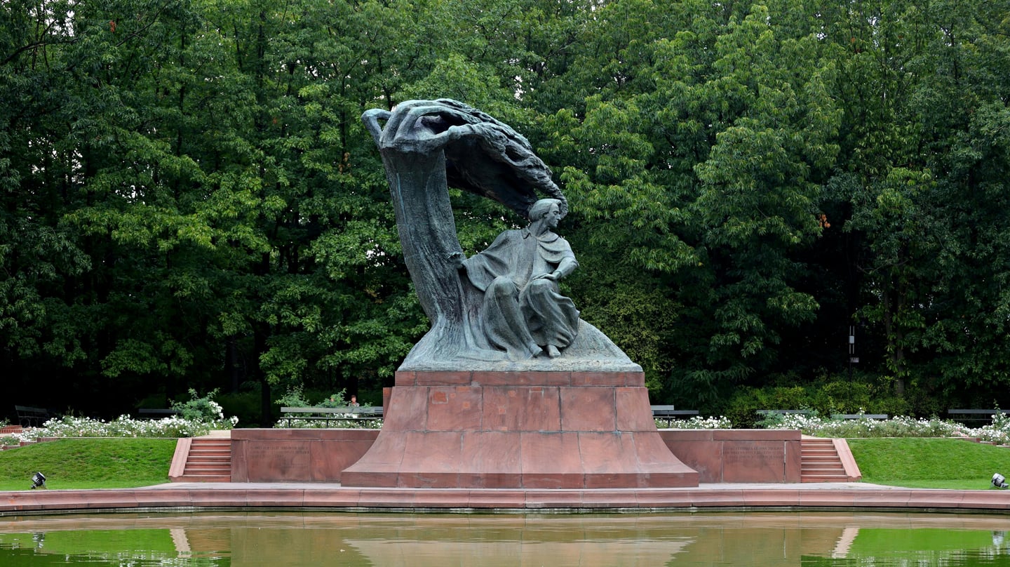 바르샤바 쇼팽 공원, 울창한 숲을 배경으로 연못 앞에 세워진 거대한 프레데리크 쇼팽 기념비와 평화로운 공원 전경입니다.