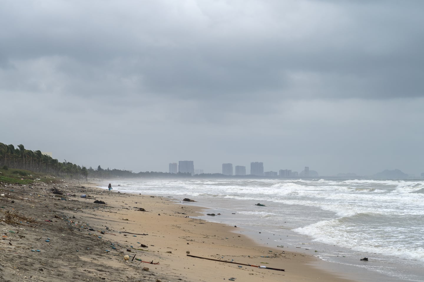 호이안 안방 비치, 먹구름 낀 하늘 아래 거친 파도가 밀려오는 해변과 멀리 보이는 도시의 실루엣이 대조를 이루는 풍경입니다.