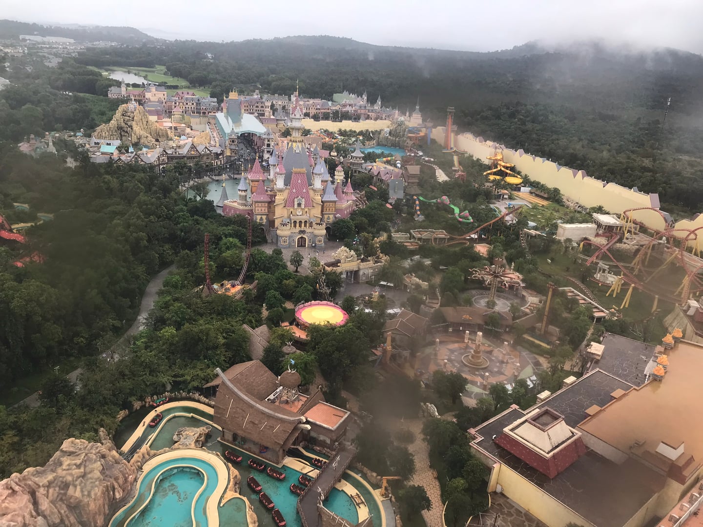 푸꾸옥 빈원더스, 안개 낀 숲속에 자리 잡은 화려한 성과 스릴 넘치는 놀이기구들이 가득한 테마파크의 웅장한 전경입니다.