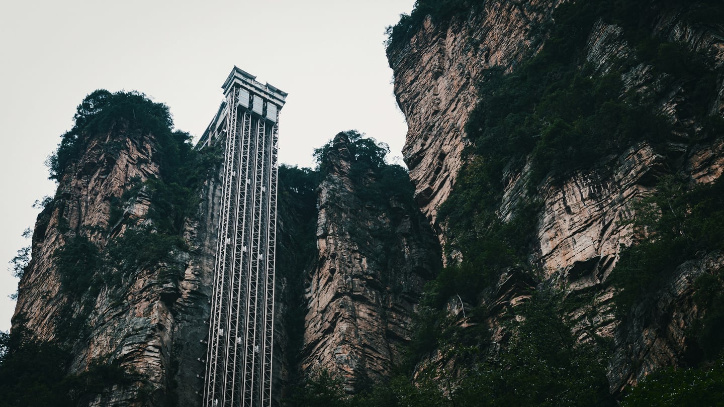 장가계 백룡 엘리베이터, 깎아지른 듯한 거대한 사암 절벽을 따라 수직으로 높게 솟아오른 웅장한 철골 구조물과 주변의 기암괴석 풍경입니다.