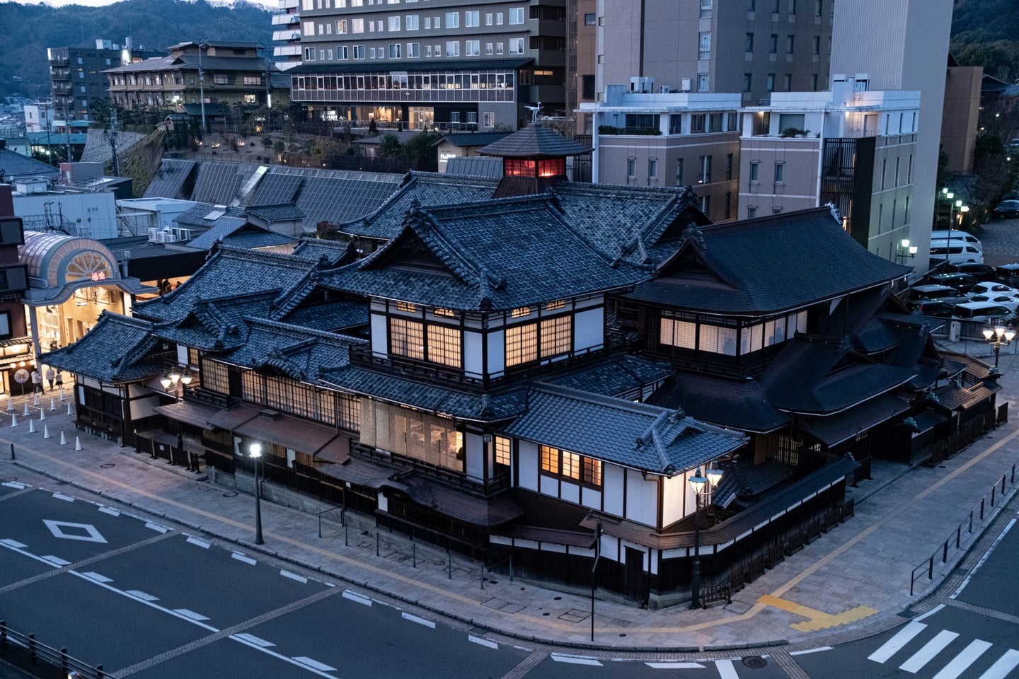 마쓰야마 도고 온천, 어스름한 저녁 빛 아래 고풍스러운 기와지붕과 창문 사이로 흐르는 따뜻한 조명이 어우러진 본관 전경입니다.
