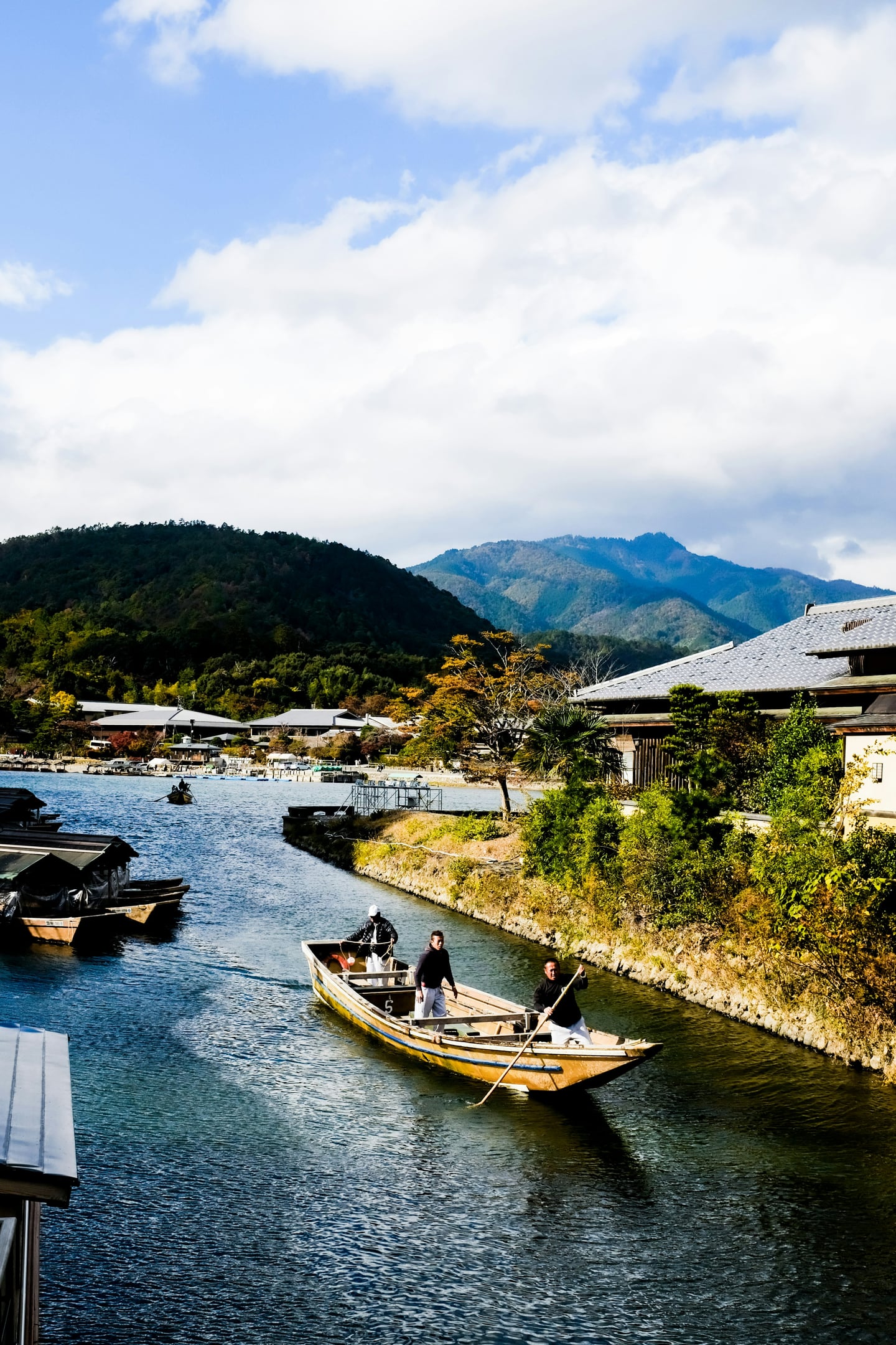 교토 도게츠교 뱃놀이, 맑은 하늘 아래 아라시야마의 푸른 산을 배경으로 가츠라 강 위를 유유히 떠가는 전통 목조 나룻배와 뱃사공들의 모습입니다.