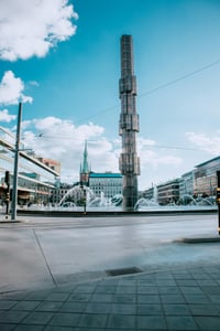 스톡홀름 세르옐스 광장, 푸른 하늘 아래 높게 솟은 유리 오벨리스크 조형물과 시원하게 뿜어져 나오는 분수가 어우러진 도심 전경입니다.