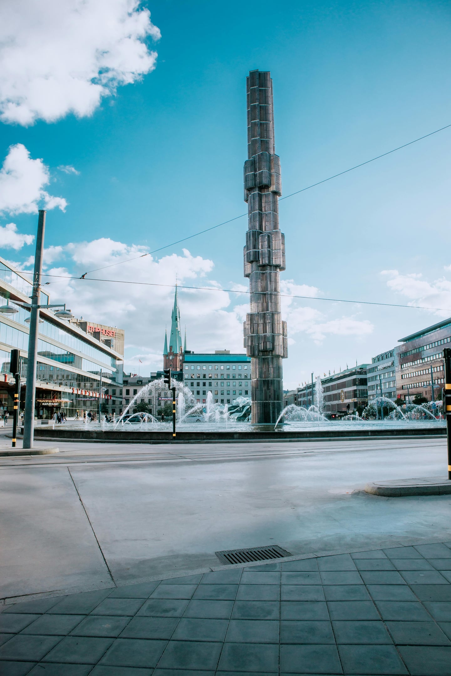 스톡홀름 세르옐스 광장, 푸른 하늘 아래 높게 솟은 유리 오벨리스크 조형물과 시원하게 뿜어져 나오는 분수가 어우러진 도심 전경입니다.