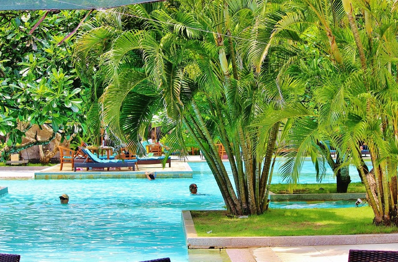 발리 누사두아, 푸른 야외 수영장을 둘러싼 울창한 야자수 아래에서 여유롭게 수영과 휴식을 즐기는 리조트의 평화로운 전경입니다.