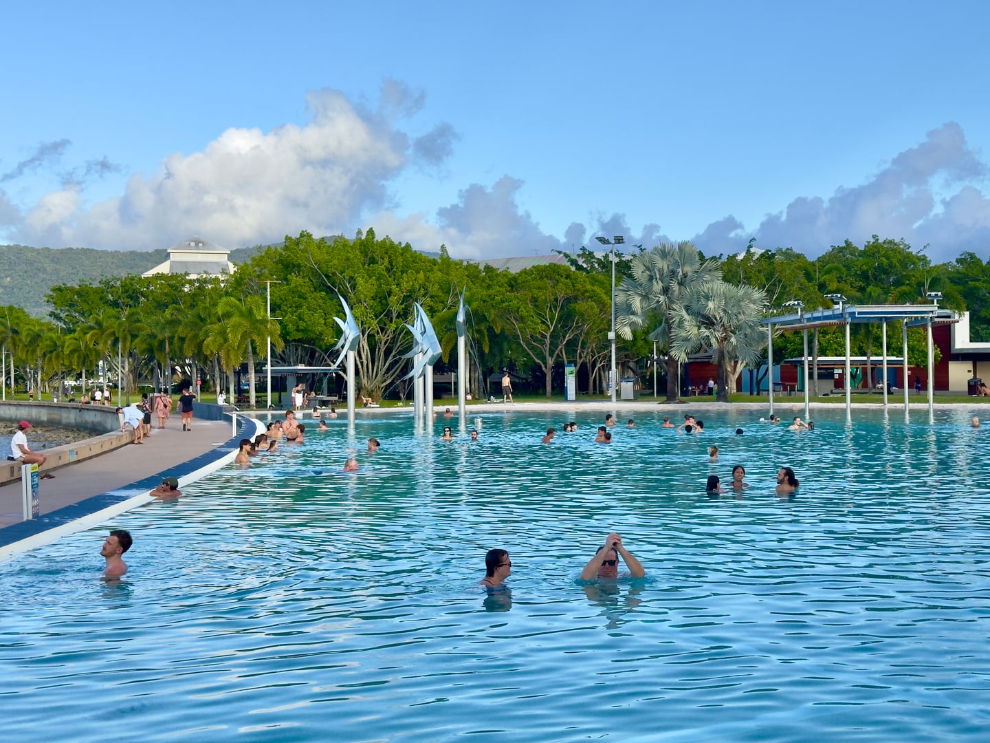 케언즈 케언즈 라군, 푸른 야외 수영장에서 물놀이를 즐기는 사람들과 상징적인 은색 물고기 조형물이 어우러진 활기찬 풍경입니다.