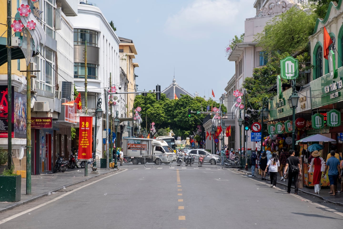 하노이 짱띠엔 거리, 프랑스풍 건축물과 짱띠엔 아이스크림 가게가 늘어선 활기찬 도로 위로 붉은 국기가 펄럭이는 풍경입니다.
