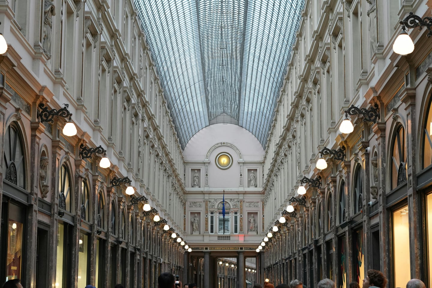 브뤼셀 생 위베르 갤러리, 웅장한 유리 천장 아래로 대칭적인 고전 건축물과 은은한 조명이 어우러진 화려한 쇼핑 거리의 모습입니다.