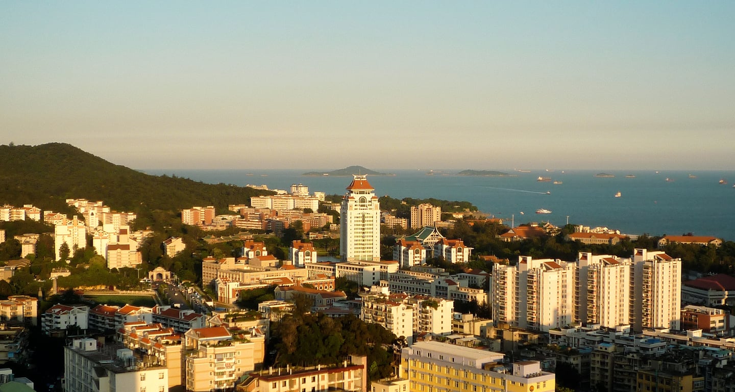 샤먼 샤먼 대학교, 푸른 바다와 산을 배경으로 붉은 지붕의 캠퍼스 건물들이 일몰 햇살 아래 펼쳐진 전경입니다.