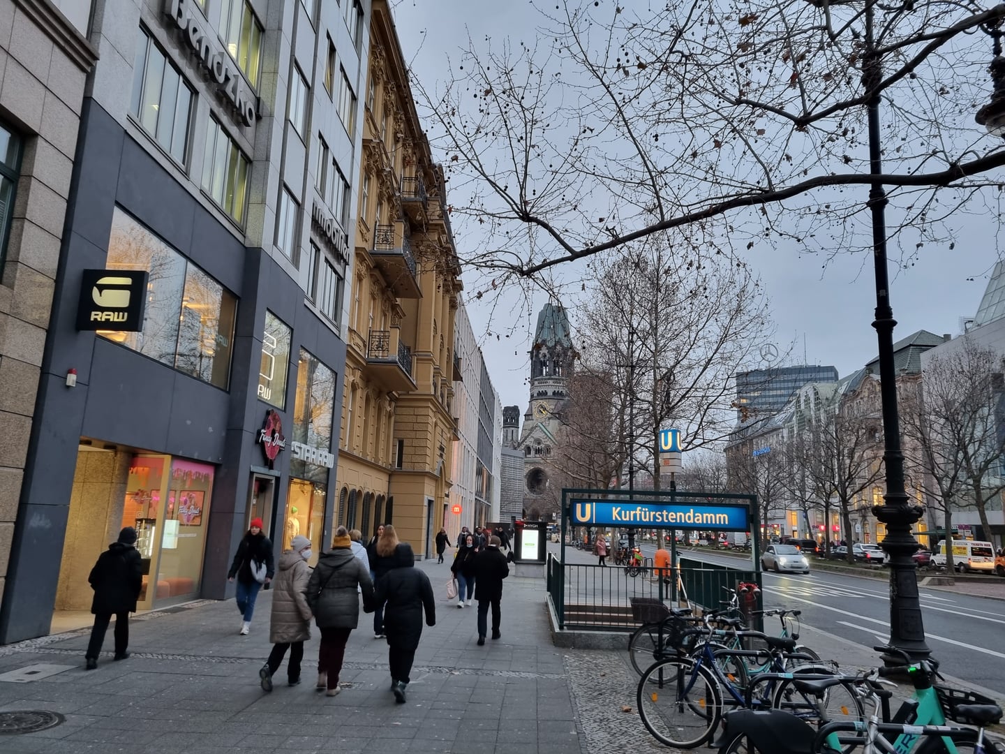 베를린 쿠담 거리, 카이저 빌헬름 기념 교회가 보이는 거리 풍경과 지하철역 입구 주변을 걷는 사람들의 모습입니다.