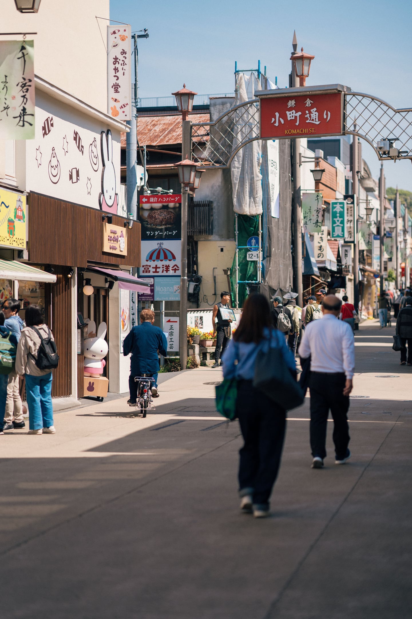 가마쿠라 고마치 거리, 붉은색 입구 표지판 아래로 미피 샵과 다양한 상점들이 늘어선 활기찬 쇼핑가의 낮 풍경입니다.