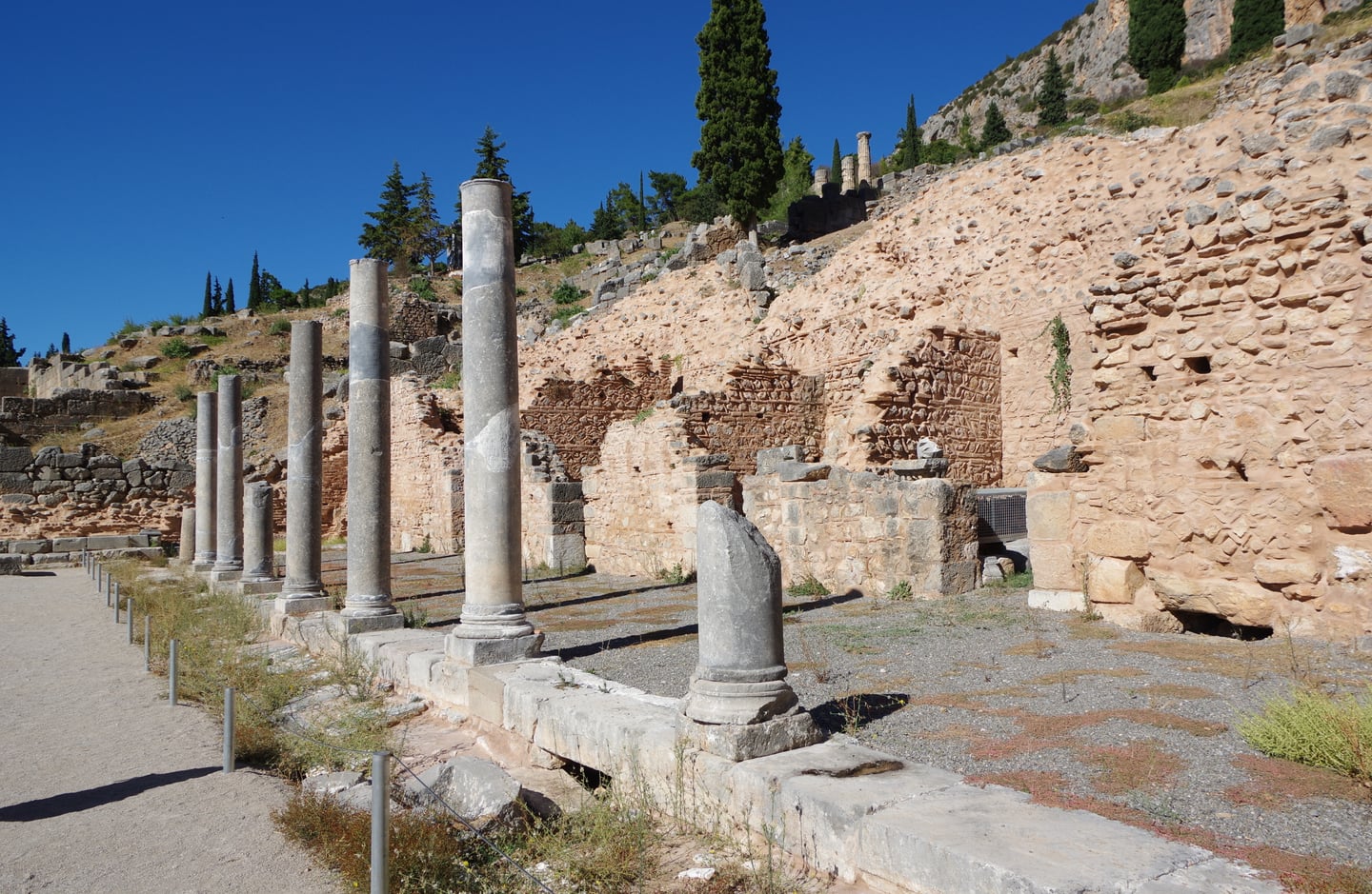 델포이 로마 아고라, 푸른 하늘 아래 일렬로 늘어선 고대 이오니아식 석조 기둥들과 세월의 흔적이 느껴지는 유적지 전경입니다.