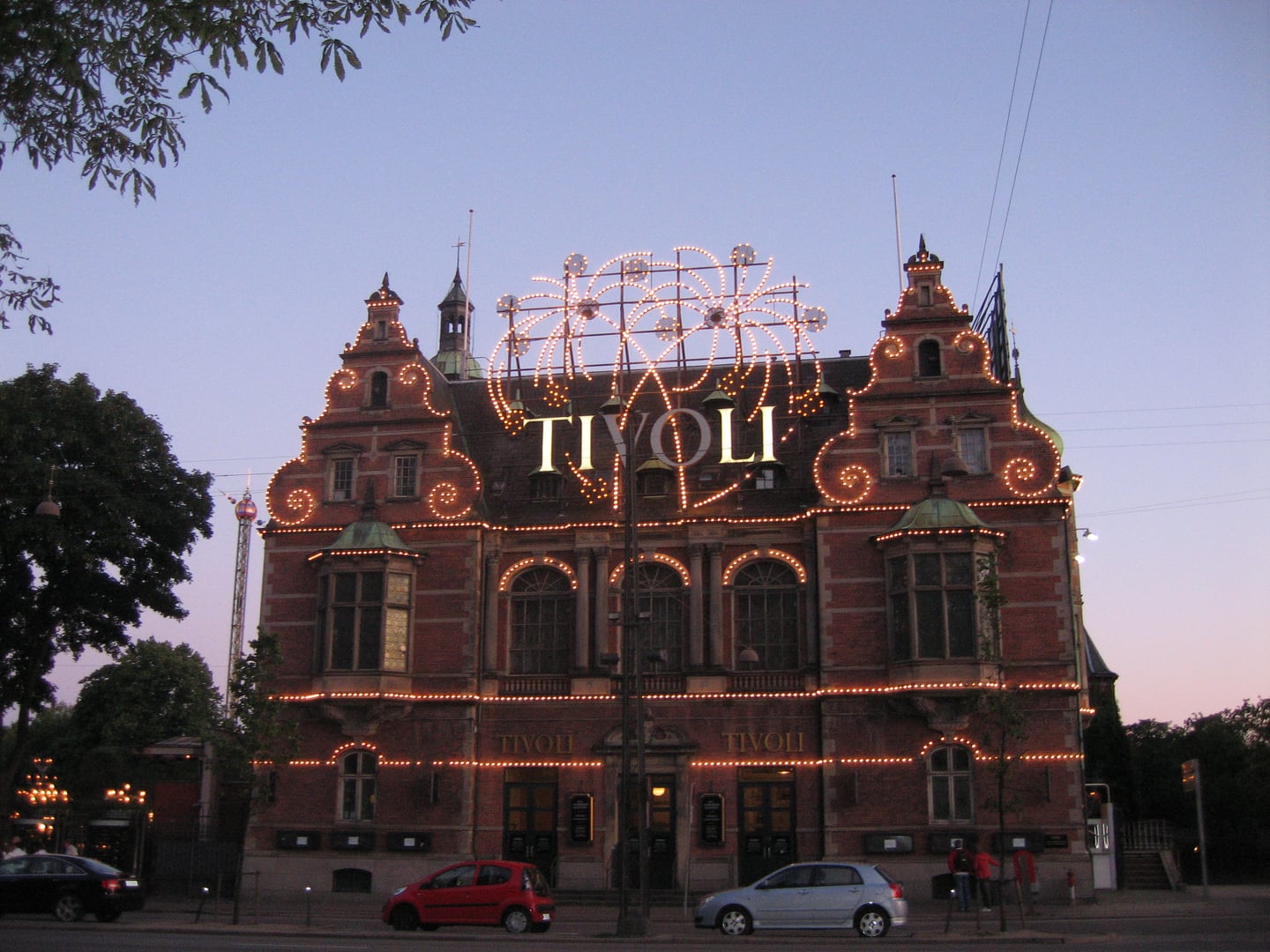 코펜하겐 티볼리 공원, 황혼의 하늘 아래 화려한 전구 장식과 네온사인이 빛나는 고풍스러운 붉은 벽돌 건물 정문의 야경입니다.