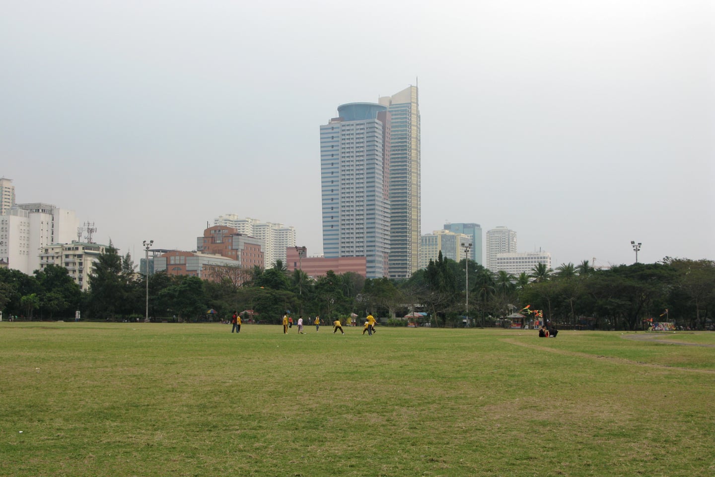 마닐라 리잘 공원, 도심 속 고층 빌딩을 배경으로 넓게 펼쳐진 푸른 잔디밭에서 여유롭게 휴식을 취하는 사람들의 풍경입니다.